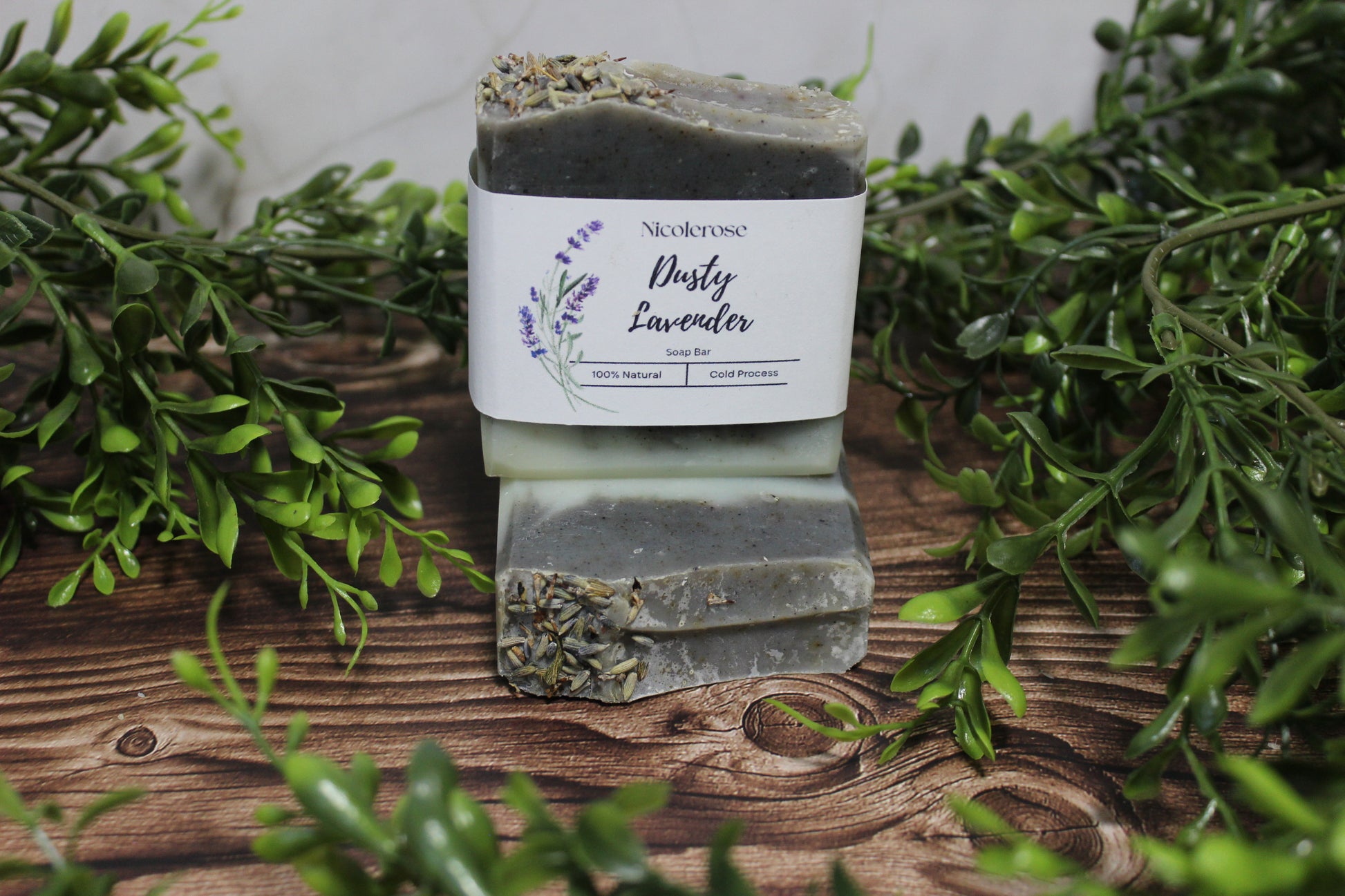 Bar of soap labeled 'Dusty Lavender' on a wooden surface with greenery