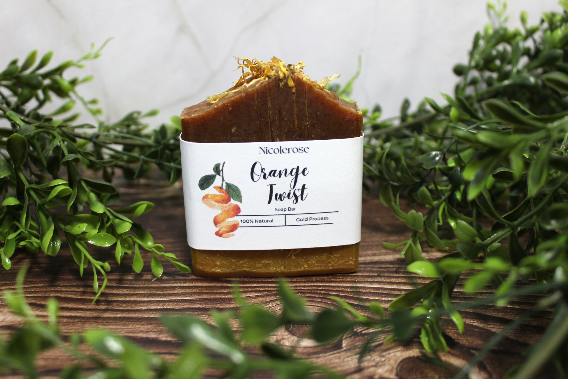 Bar of soap with 'Orange Twist' label on a wooden surface surrounded by greenery