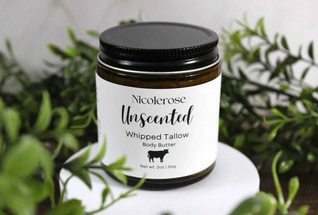 Jar of Nicoleros Unscented Whipped Tallow Body Butter with a black lid on a white surface surrounded by green plants.