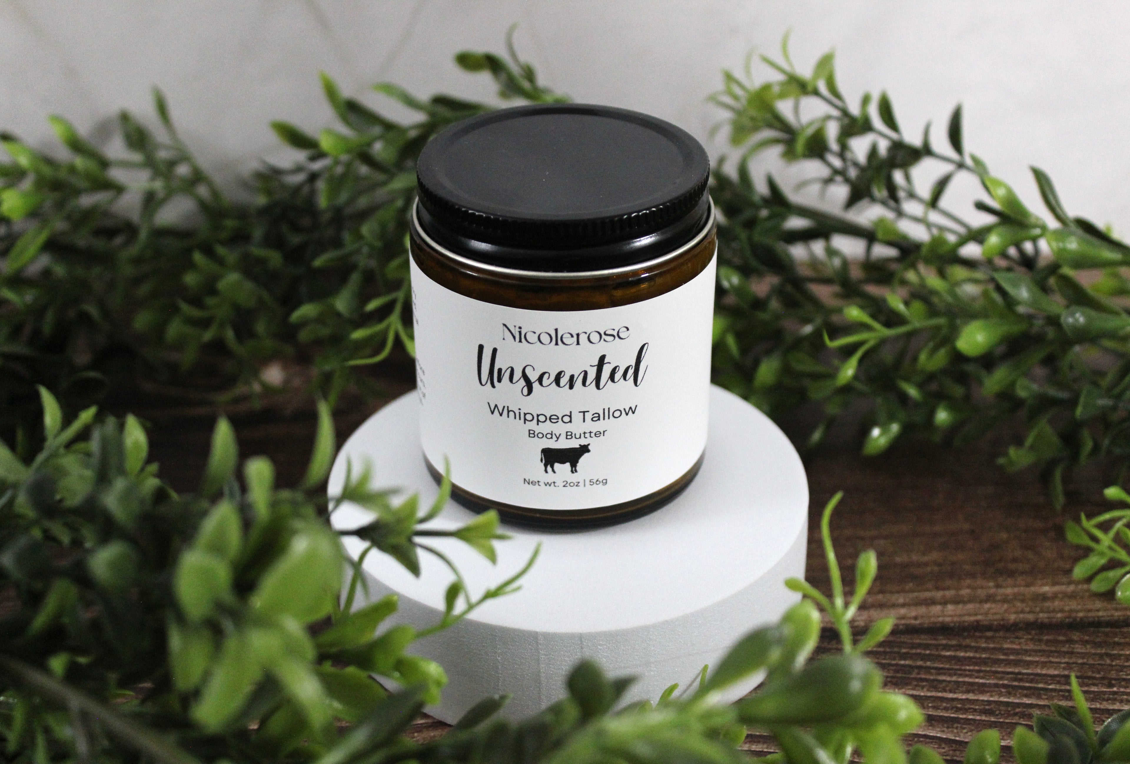 Jar of Nicolerosse Unscented Whipped Tailgall body butter on a white stand with greenery in the background