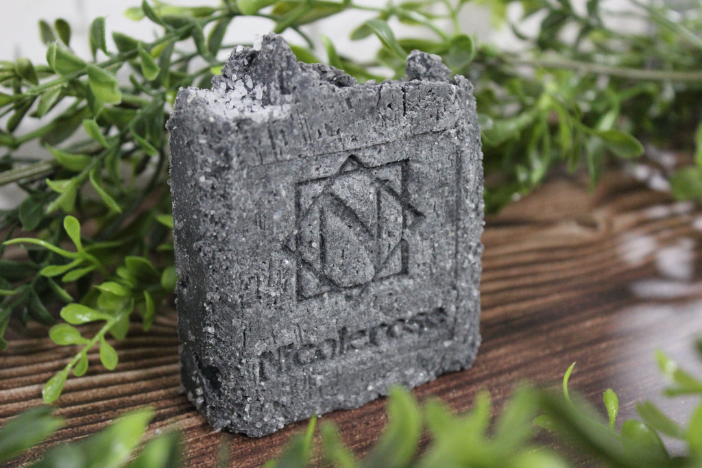 Gray textured soap bar with a logo and text on a wooden surface with green plants in the background