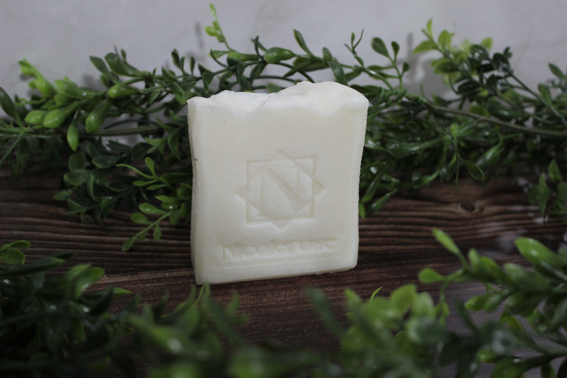 White soap bar with a logo on a wooden surface surrounded by green leaves