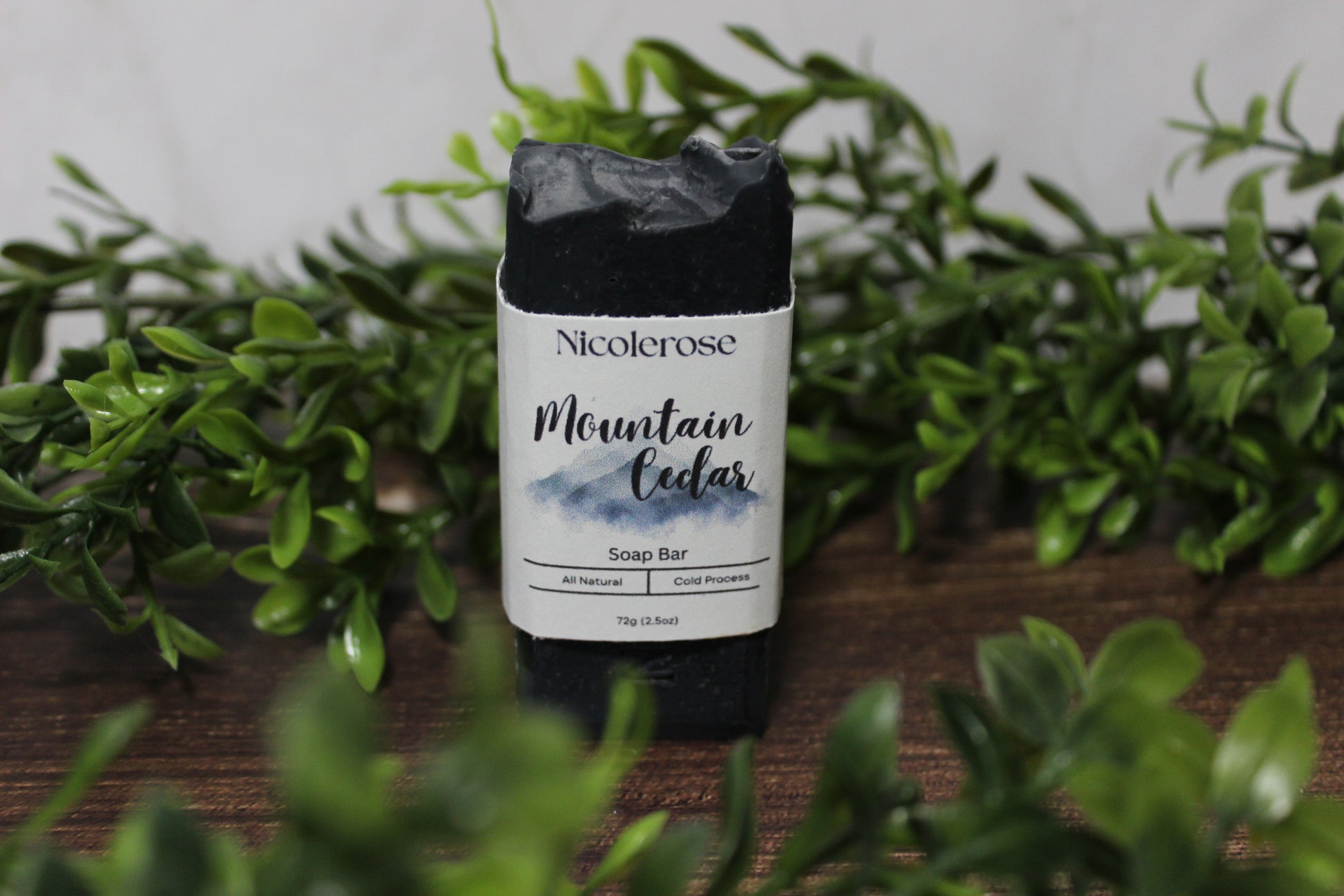 Nicolerose Mountain Cedar soap bar on a wooden surface with greenery in the background